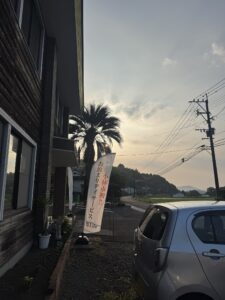 夕暮れどき、虹Lifeの建物前に立つ「小林市初!!お泊まりデイサービス」ののぼりとフェニックスの木